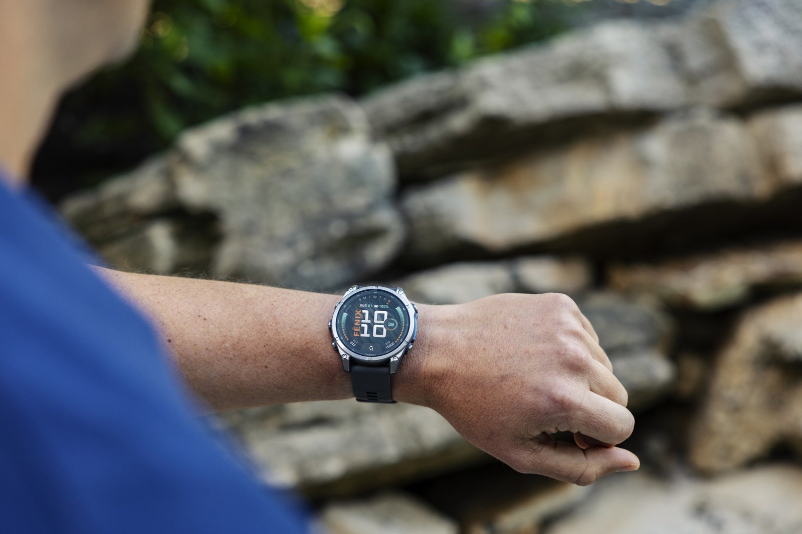 Montre Garmin fēnix 9 au poignet devant un muret de pierres, cadran de la montre allumé