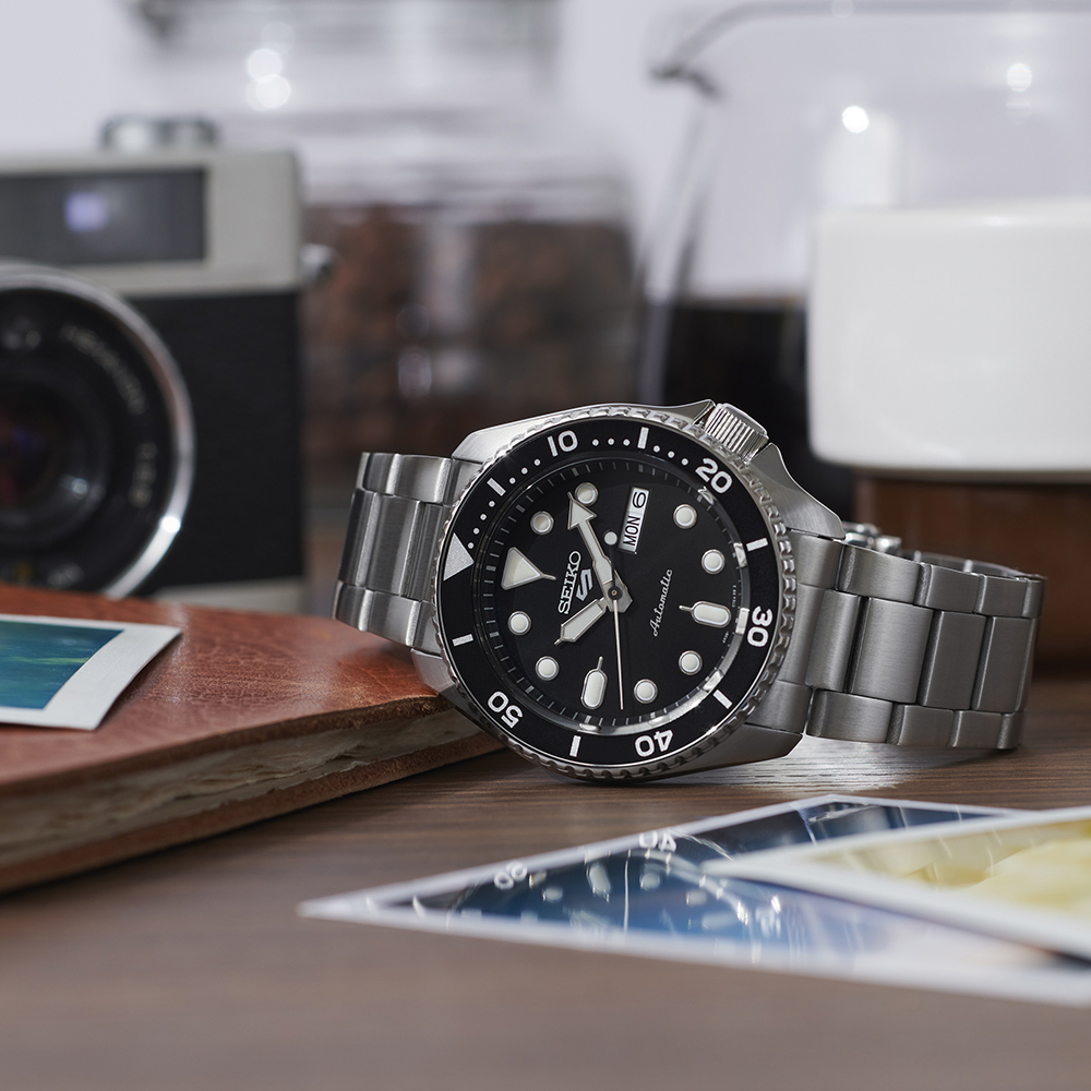 Montre Seiko 5 Sports SRPD55K1 automatique 42 mm, cadran noir, jour et date, bracelet acier, posée sur un trottoir mouillé au matin