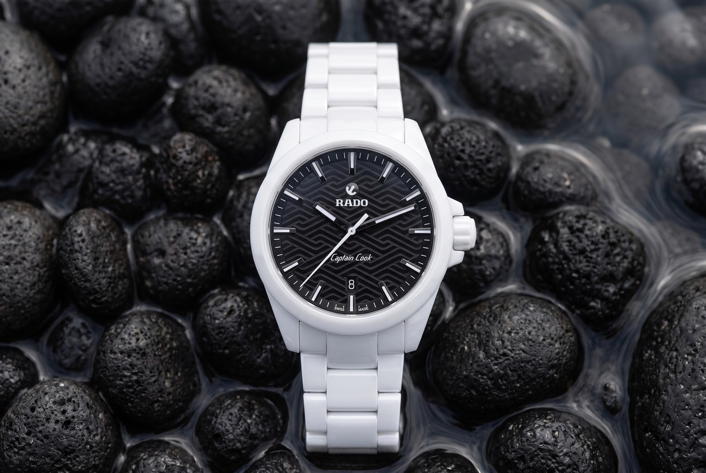 Rado Captain Cook Discover en céramique blanche, cadran noir à motif de vagues, posée sur des galets volcaniques noirs et mouillés